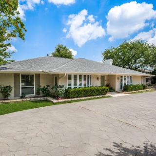 A single-story residential house with a spacious driveway, surrounded by trees and set against a blue sky with fluffy clouds.