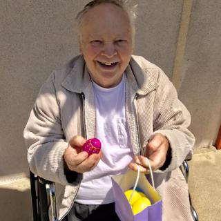 An elderly man smiling while holding a colorful egg and a gift bag outdoors.