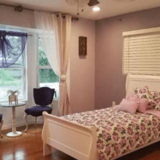 A cozy bedroom with purple walls, featuring a single bed adorned with decorative pillows and a bay window with white curtains.