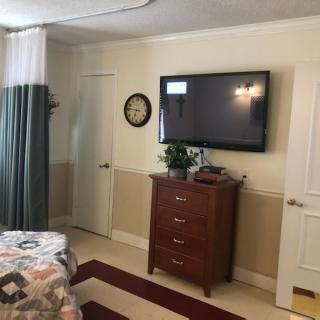 Cozy bedroom with a flat-screen TV mounted on the wall, a dresser below, and a bed with patterned bedding. The room features curtains and a decorative plant on the dresser.