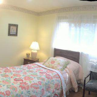 A cozy bedroom with a single bed covered in floral bedding, a bedside lamp, and a window letting in natural light.