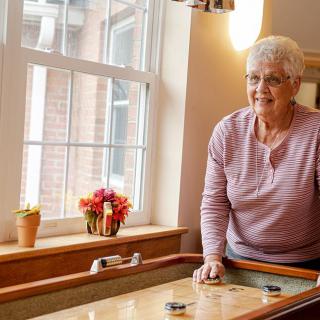 An elderly woman enjoys a game of shuffleboard indoors by the window, with flowers in the background.