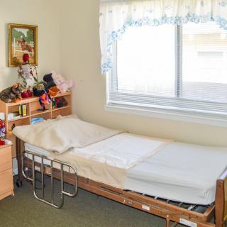 A cozy bedroom featuring a hospital bed, a window with lace curtains, and a neatly organized bookshelf filled with books and decorations.