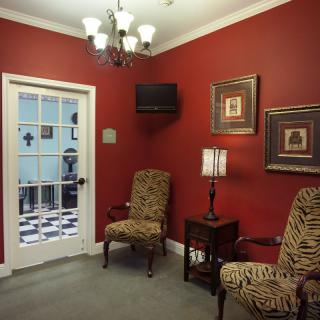 A cozy waiting room with red walls, featuring two patterned chairs, a small table with a lamp, and wall art.