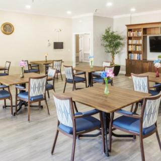 A modern dining area with wooden tables and chairs, a bookcase, indoor plant, and bright lighting.