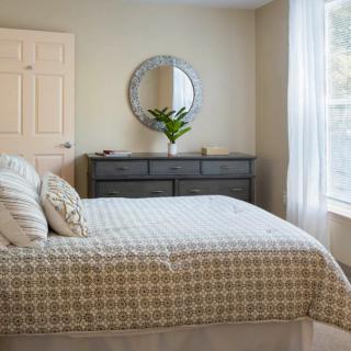 A cozy bedroom with a neatly made bed, dresser, and round mirror by the window.