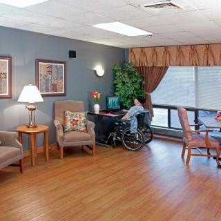 A cozy lounge area in a nursing home with comfortable chairs, a wheelchair user by a desk, and large windows letting in natural light.
