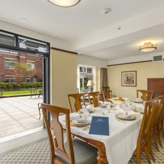 A well-lit dining room with a table set for a meal, featuring wooden chairs and windows overlooking a garden.