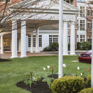 A building entrance with white columns, a parked car, and landscaped greenery.