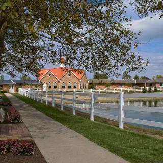 A scenic view of a building with a red roof, surrounded by a pond, pathway, and white fence under a partly cloudy sky.