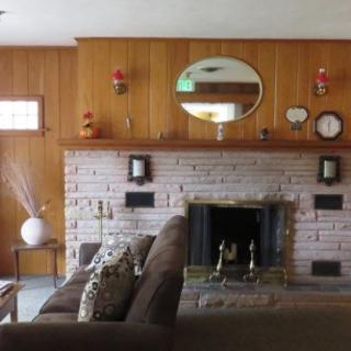 A cozy living room featuring a brick fireplace and wood-paneled walls, with a large mirror above the mantle.