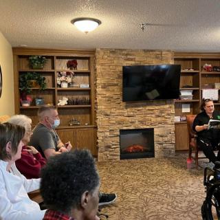 A group of people sitting in a cozy room with bookshelves and a fireplace, listening to a speaker.