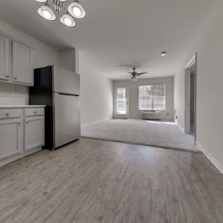 A modern apartment interior featuring a kitchen area with cabinets, fridge, and wood flooring adjacent to a living space with large windows and a ceiling fan.