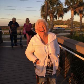An elderly woman smiling joyfully while walking on a boardwalk during sunset.