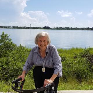 An elderly woman enjoys a sunny day by the lake, using a walking aid.