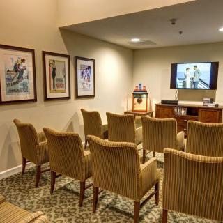 A quiet waiting room with rows of upholstered chairs facing a television on the wall, adorned with framed artwork and a patterned carpet.