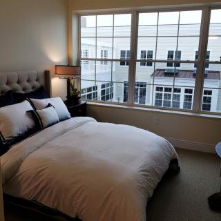 A cozy bedroom with a neatly made bed and large windows overlooking neighboring buildings.
