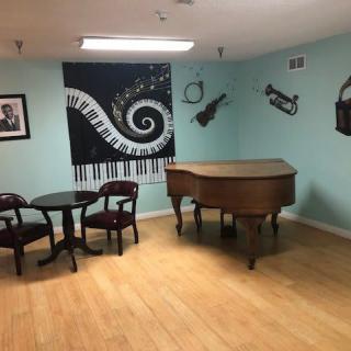 A cozy music-themed room featuring a grand piano, decorative wall art of musical instruments, and a small table with chairs on a hardwood floor.