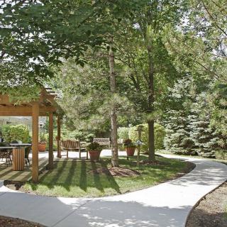 A tranquil garden with a winding pathway, shaded by trees and featuring a pergola.
