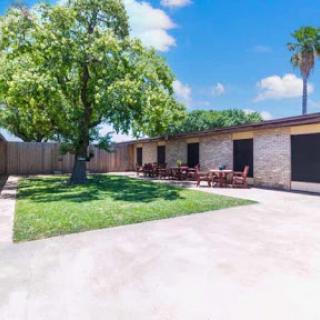 A sunny outdoor view of a home featuring a spacious garden, a large tree, and patio furniture.