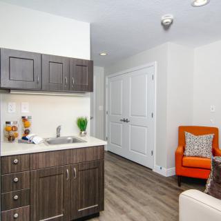 A modern kitchenette with dark cabinetry and an orange chair in the corner of a living space.