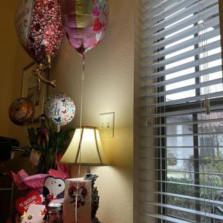 A cozy room corner with celebratory balloons, a bouquet of flowers, and a decorative lamp by the window.