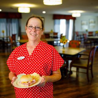 A smiling woman in a red polka dot shirt holds a plate inside a dining area.