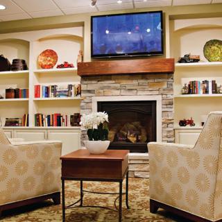A cozy living room with a fireplace, flanked by bookshelves, two armchairs, and a television mounted above.