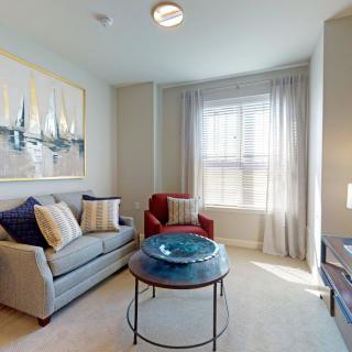 A cozy living room with a sofa and armchair, featuring a large window with curtains and a decorative painting on the wall.