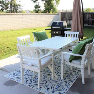 A backyard patio setup featuring a white table with chairs, a grill, and an umbrella on a patterned rug.