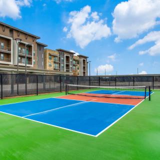 An outdoor tennis court with vibrant blue and green surfaces, surrounded by fencing and apartment buildings under a clear blue sky.