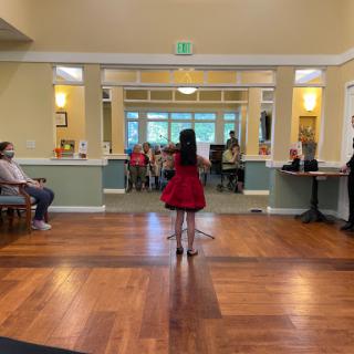 A young girl performs on stage in a red dress while an audience looks on, showcasing talent and joy at a community gathering.