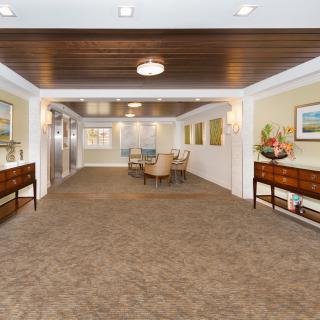 A spacious and elegantly decorated lobby area featuring a carpeted floor, wooden ceiling with lights, and furnishings including tables and chairs.