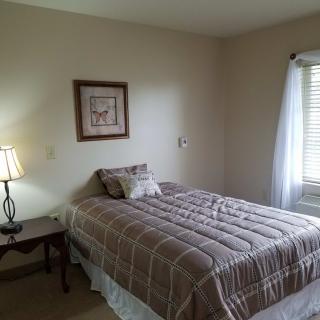 A cozy bedroom with a neatly made bed, a lamp on a side table, and a window letting in natural light.