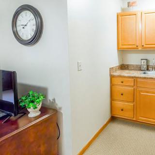 A small kitchenette area featuring wooden cabinets, a sink, a wall clock, and a nearby TV with a decorative plant.