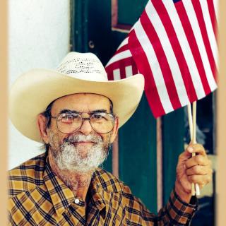 A smiling elderly man with a beard and glasses holds an American flag while wearing a cowboy hat and a plaid shirt.