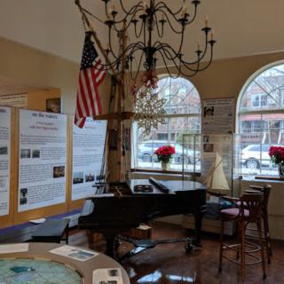 A room with a piano, chandelier, American flag, and informational posters by large arched windows.