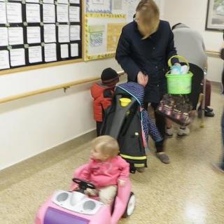 A caregiver with children in a hallway, pushing a stroller while one child rides a toy car.