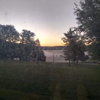 A serene view of a misty morning by the water, with trees framing the horizon as the sun begins to rise.