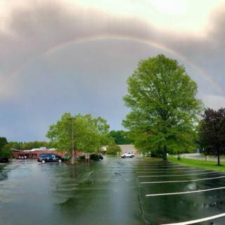 A serene rainbow arches over a quiet parking lot surrounded by lush green trees.