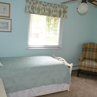 A cozy bedroom with a single bed and armchair, featuring light blue walls and a window bringing in natural light.