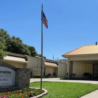 A building with an American flag in front, featuring a sign and surrounded by greenery under a clear blue sky.
