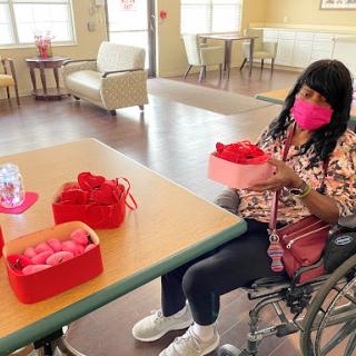 A woman in a wheelchair enjoys a crafting activity with red decorations and gifts in a community center.