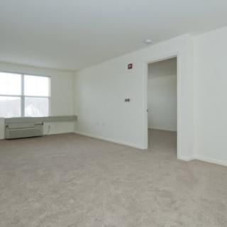 An empty room with white walls, a large window, and carpeted flooring.