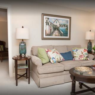 A cozy living room with a beige sofa, colorful cushions, two side tables with lamps, and a framed painting on the wall.