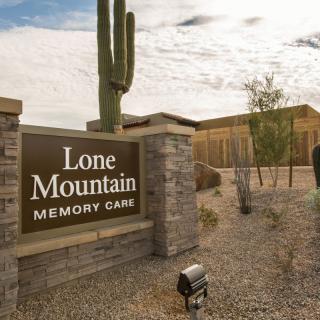 Entrance sign for Lone Mountain Memory Care set against a desert backdrop with a cactus and surrounding buildings.