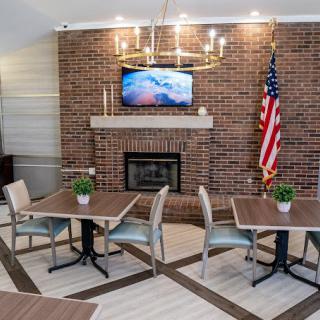 A cozy dining area with a brick fireplace, featuring modern tables and chairs, a TV mounted above the mantel, a chandelier overhead, and an American flag displayed in the corner.
