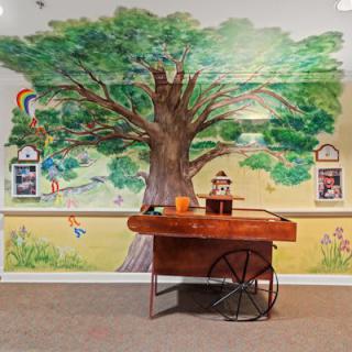 A colorful indoor mural featuring a large tree, with a wooden cart in front of it.