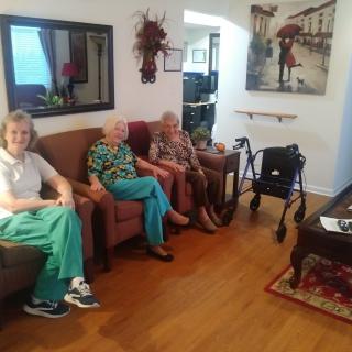 Three elderly women sitting in a cozy living room, with a walker nearby and artwork on the wall.