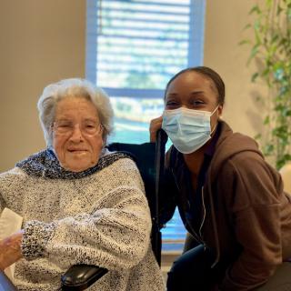 An elderly woman sitting in a wheelchair, smiling alongside a caretaker wearing a mask indoors.